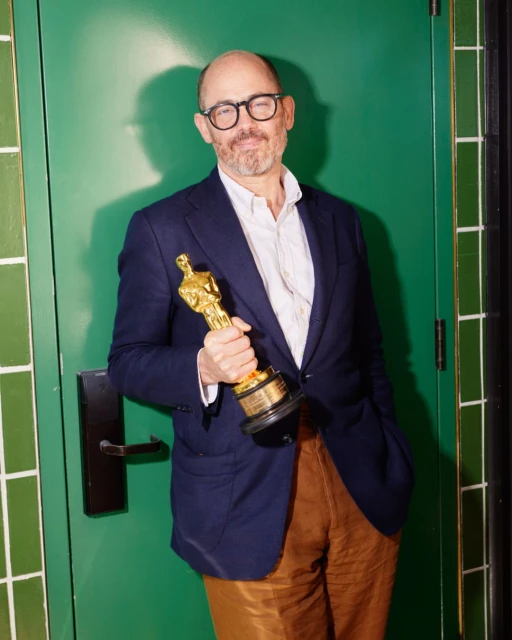 Adult male poses with a gold-colored statuette in front of a green door with white and green tiling in a medium indoor shot.