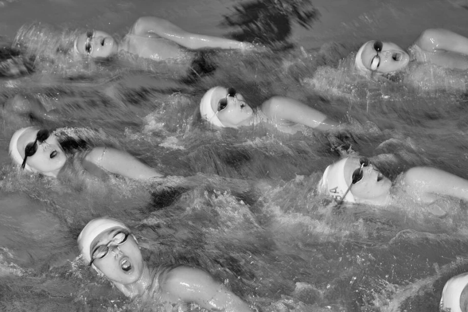 Black and white photo shows multiple swimmers in a pool wearing swim caps and goggles, competing in a swimming event with splashes.
