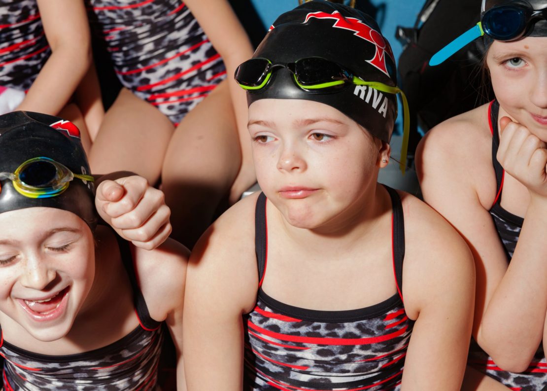 Three young female athletes in swimsuits and caps gather indoors, smiling and showing varied facial expressions; group portrait, swimwear.