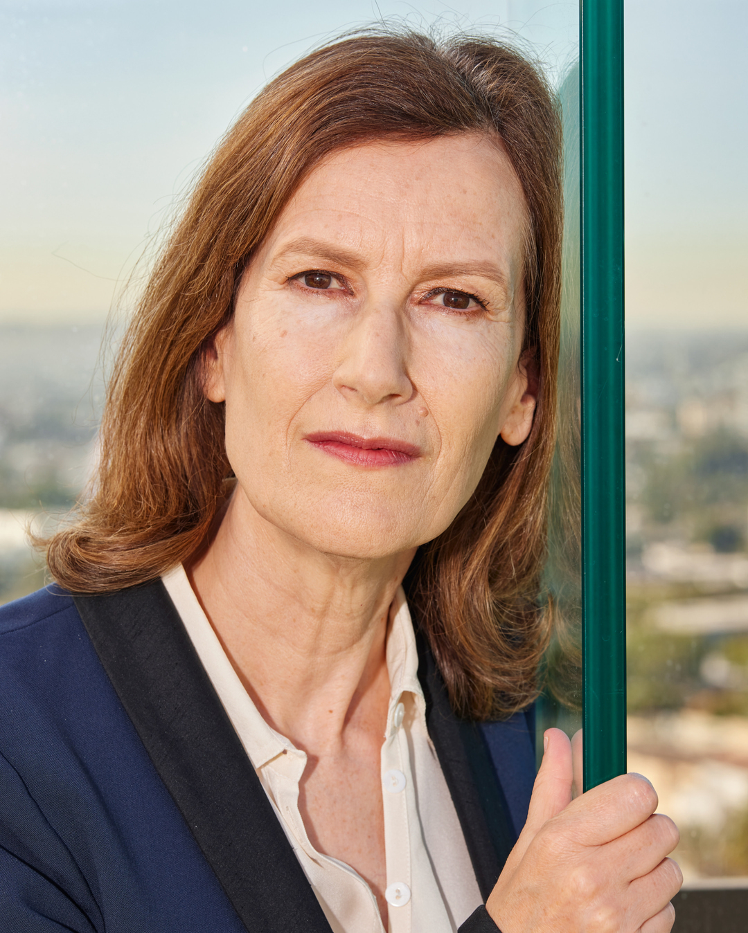 Portrait of an adult woman with auburn hair, wearing a navy jacket and cream-colored blouse, posing indoors with a blurred background.