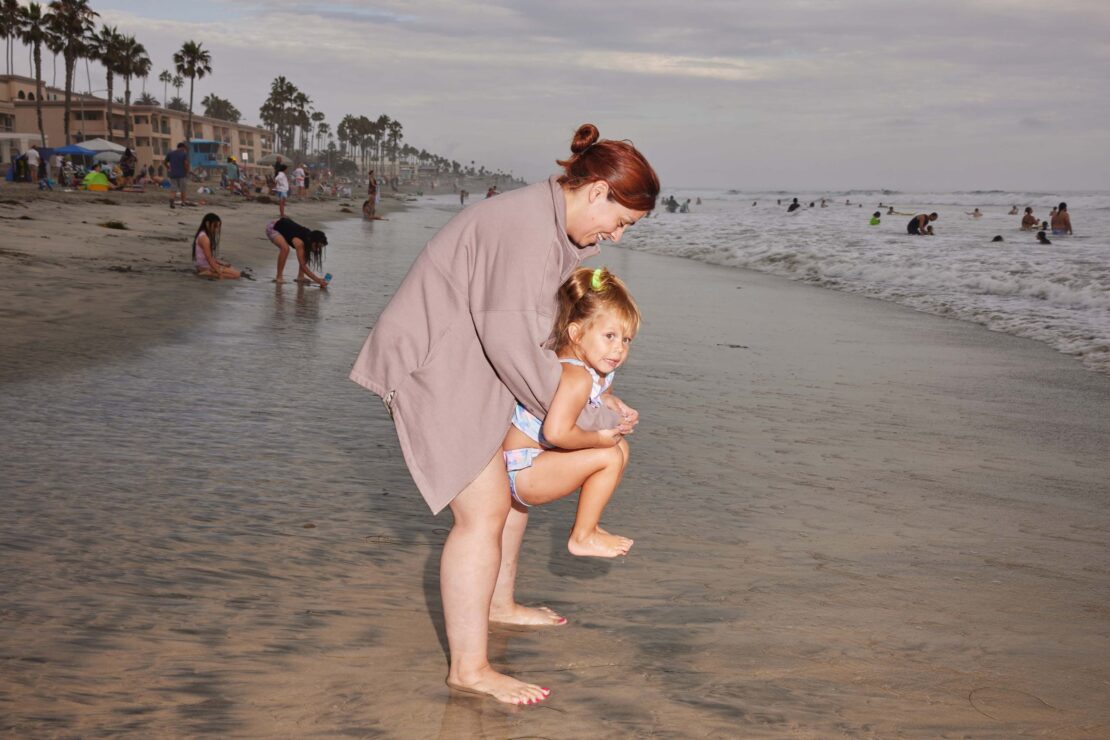 Woman holding a young child on a sandy beach near the ocean by Maggie Shannon
