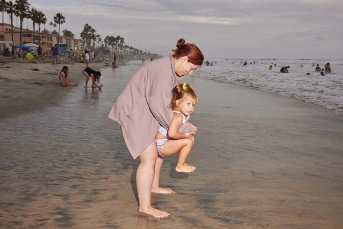 Woman holding a young child on a sandy beach near the ocean by Maggie Shannon