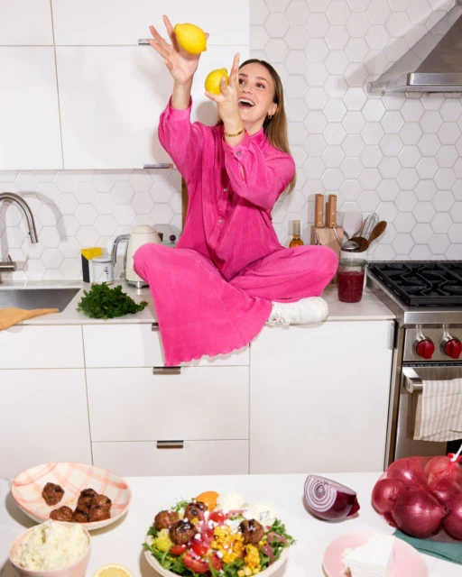 A woman in a pink jumpsuit sits cross-legged on a white kitchen counter, juggling lemons with a bright salad and red onions nearby.