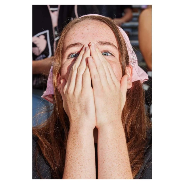 A close-up of a young adult woman with long auburn hair wearing a pink bandana covering her mouth and nose with both hands.