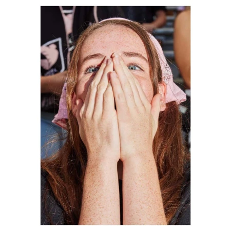 A close-up of a young adult woman with long auburn hair wearing a pink bandana covering her mouth and nose with both hands.