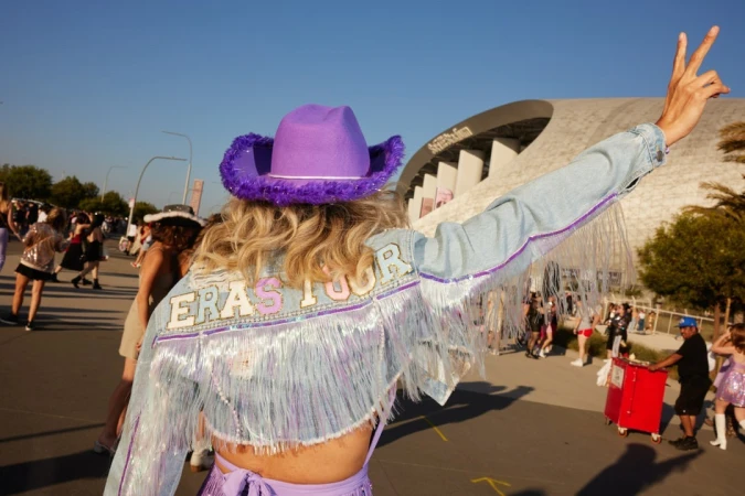 Crowd scene with a woman facing away wearing a denim jacket with fringe and a purple cowboy hat outside of SoFi Stadium.