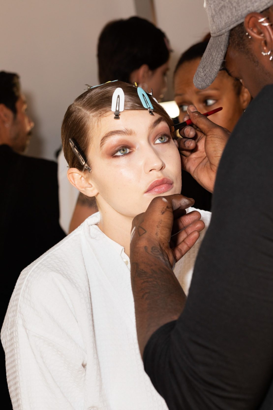 Model getting makeup applied backstage, wearing a white robe, hair clipped back, makeup artist standing at the side applying makeup.