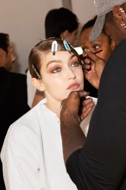 Model getting makeup applied backstage, wearing a white robe, hair clipped back, makeup artist standing at the side applying makeup.