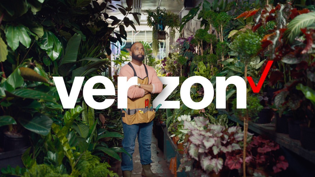 Man stands in indoor plant shop filled with greenery. He wears a tan apron over a pink shirt. Verizon logo is prominent in foreground.