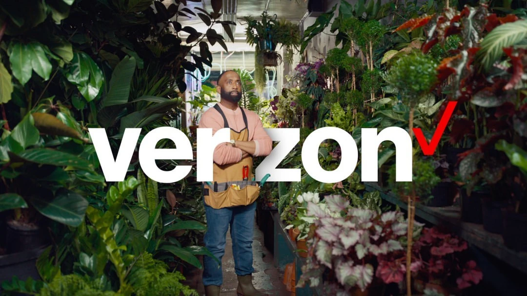 Man stands in indoor plant shop filled with greenery. He wears a tan apron over a pink shirt. Verizon logo is prominent in foreground.