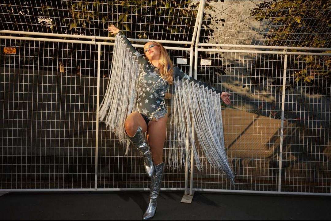 A woman in a star-patterned bodysuit and fringed sleeves poses in silver boots in front of a chain-link fence outdoors.