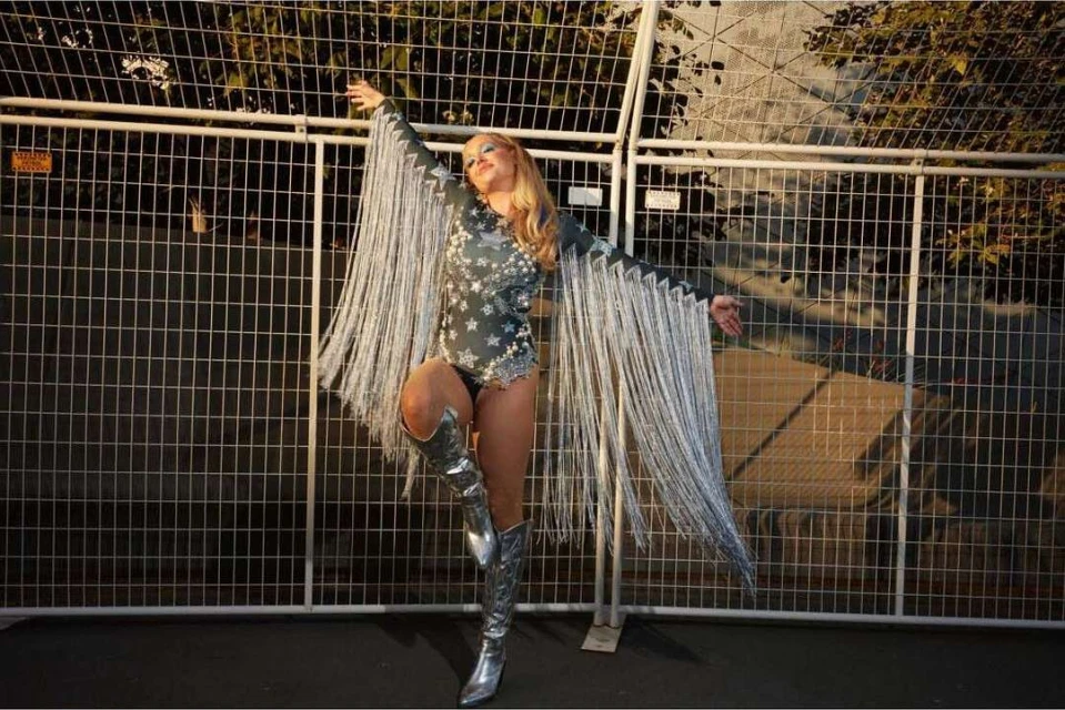 A woman in a star-patterned bodysuit and fringed sleeves poses in silver boots in front of a chain-link fence outdoors.