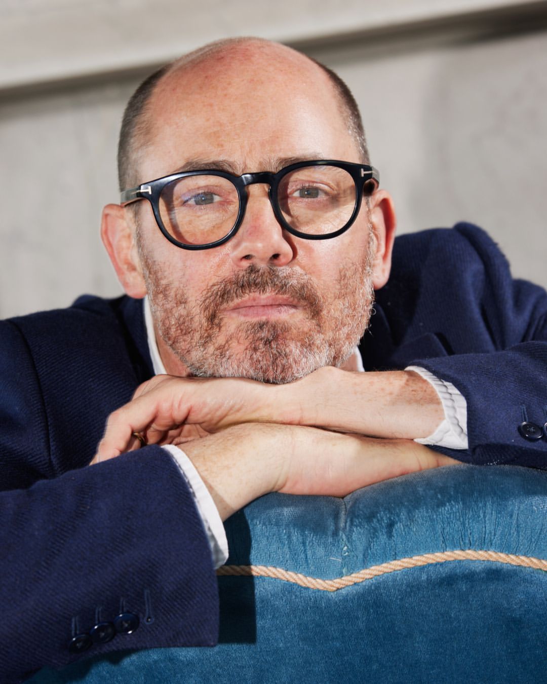 Close-up portrait of a mature adult man with eyeglasses wearing a navy blue jacket, leaning his arms on a teal-colored seat cushion indoors.