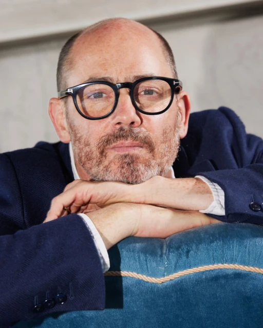 Close-up portrait of a mature adult man with eyeglasses wearing a navy blue jacket, leaning his arms on a teal-colored seat cushion indoors.