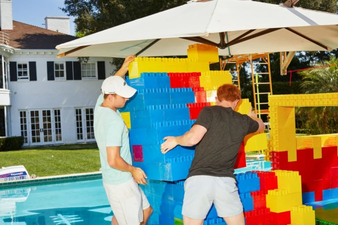Two men build a large colorful structure from toy blocks outdoors next to a swimming pool, with a large white house in the background.