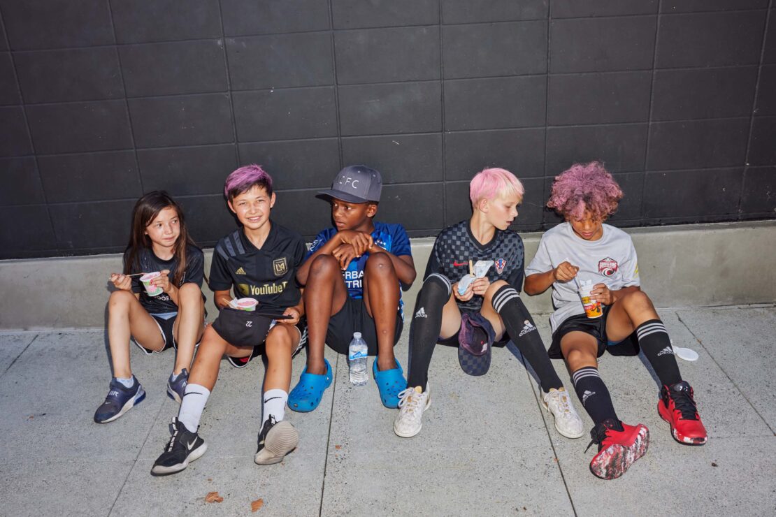 Five kids in sports uniforms sit against a dark wall and eat. Three have dyed hair in pink or purple hues and wear soccer attire.