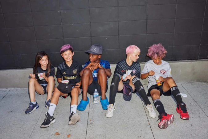 Five kids in sports uniforms sit against a dark wall and eat. Three have dyed hair in pink or purple hues and wear soccer attire.
