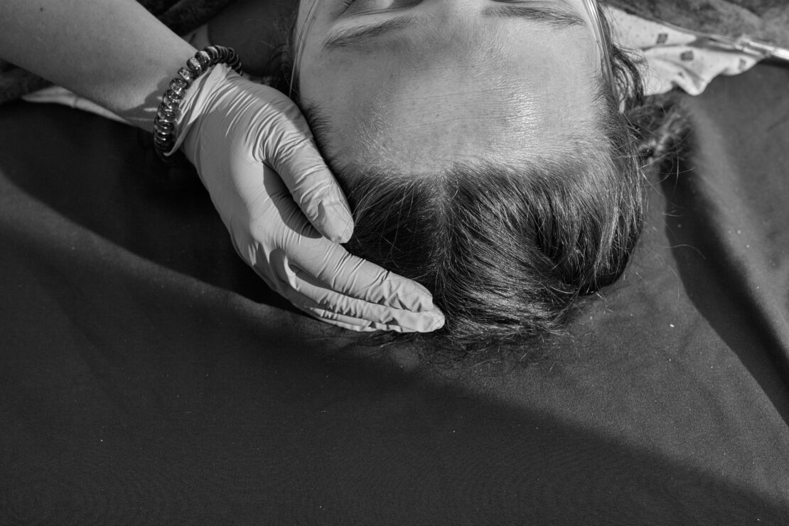 Black and white photograph of an adult with a hand in a glove touching their head. The scene is set on a dark surface.