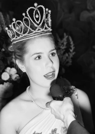 Monochrome portrait of a young woman wearing an elaborate pearl-encrusted tiara while speaking into a handheld microphone.