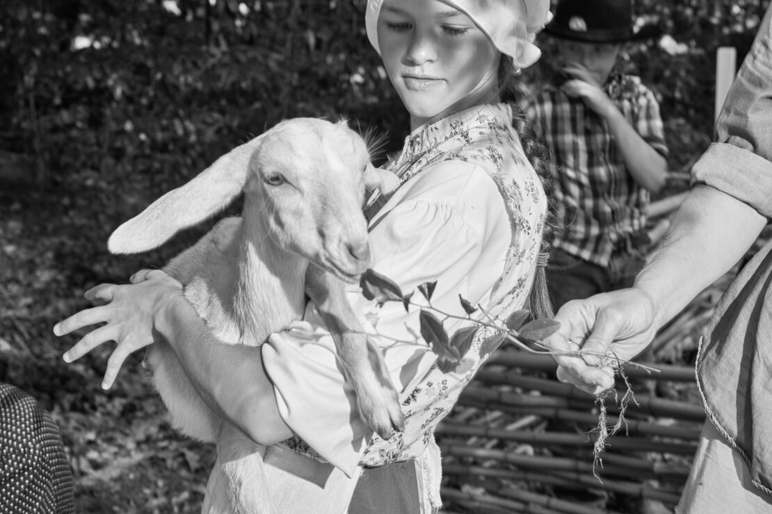 Young girl in historical clothing holds a white goat in a monochrome outdoor setting, with another child visible in the background.