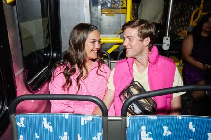 Smiling couple riding bus: Woman in pink textured shirt, man in pink vest, engaging in conversation on blue patterned seats.