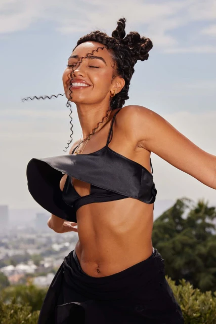 A smiling woman with curly updo is captured in a medium shot against a bright outdoor backdrop with city skyline and trees visible.