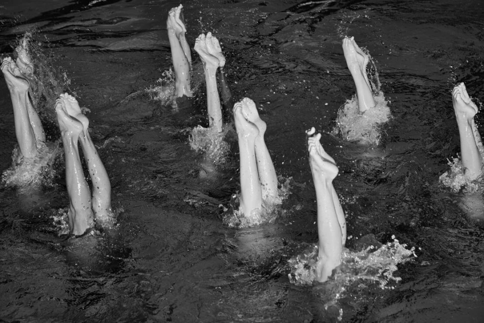 Monochrome view of synchronized swimmers with legs and feet extended upward from the water, creating dynamic splash patterns and movement.
