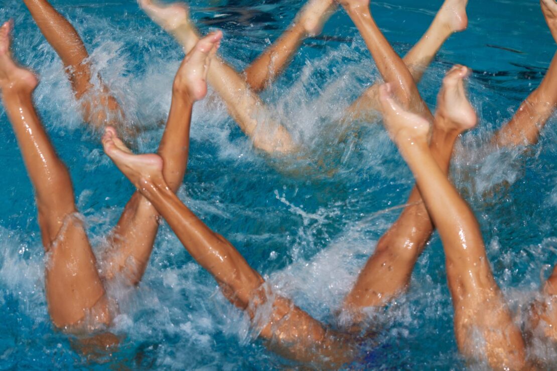 Synchronized swimming performance featuring multiple swimmers' legs and feet extending out of the blue water in a choreographed formation.