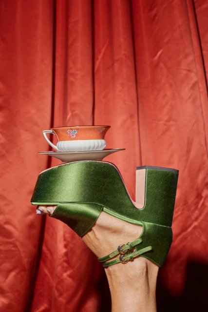 Elevated view of a green platform shoe on a woman's foot with a decorative teacup on top, set against a draped red fabric background.