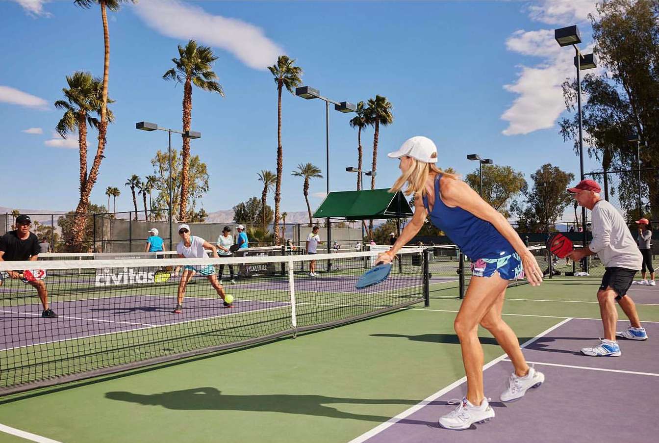 A woman hits a pickleball in an outdoor game; other players and palm trees are visible on a sunny day at the pickleball court.