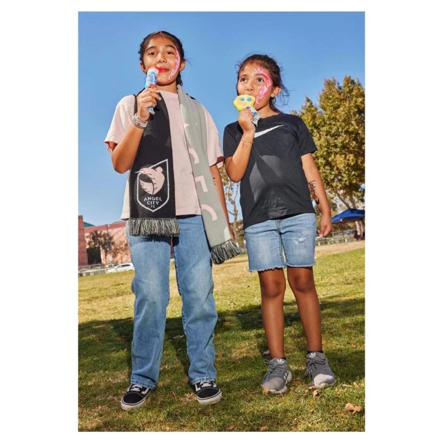Two young girls with face paint stand on a grassy field holding lollipops, with a soccer scarf, buildings, and blue sky in the background.
