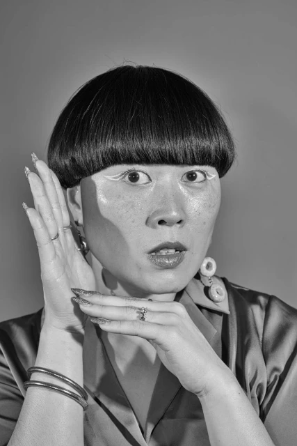Black and white portrait of an adult woman with bowl cut and statement makeup holding her hands near her face in an indoor setting.