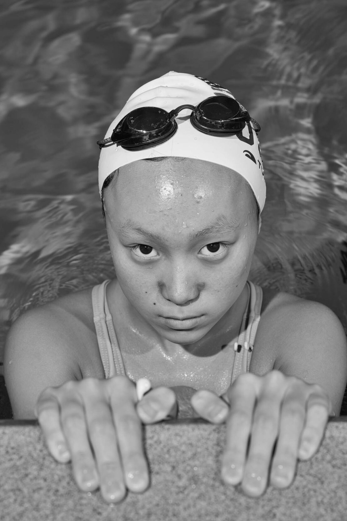 Asian female swimmer in cap and goggles holding onto the pool edge with water background in black and white photograph. 