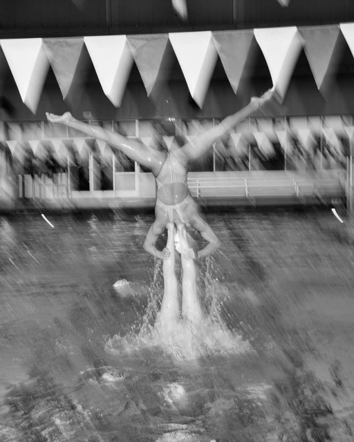 Monochrome photo of two synchronized swimmers performing a complex lift in a pool, creating a dynamic splash with decorative triangular pennants.
