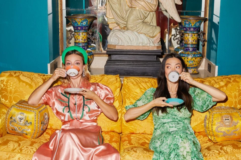 Two adult women sit on a yellow patterned couch, drinking from teacups and saucers, in an indoor setting with decorative objects.