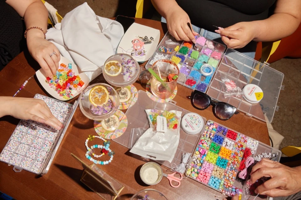 Overhead shot of four sets of hands crafting beaded bracelets at a wooden table surrounded by bead containers and beverage glasses.