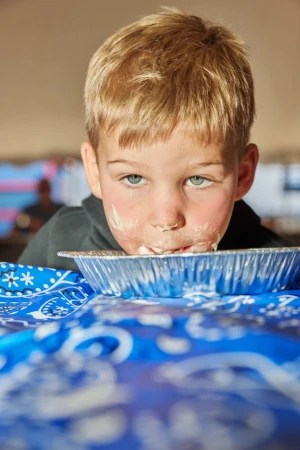 Young child with messy face leaning over a disposable foil pie tin resting on a blue patterned table runner