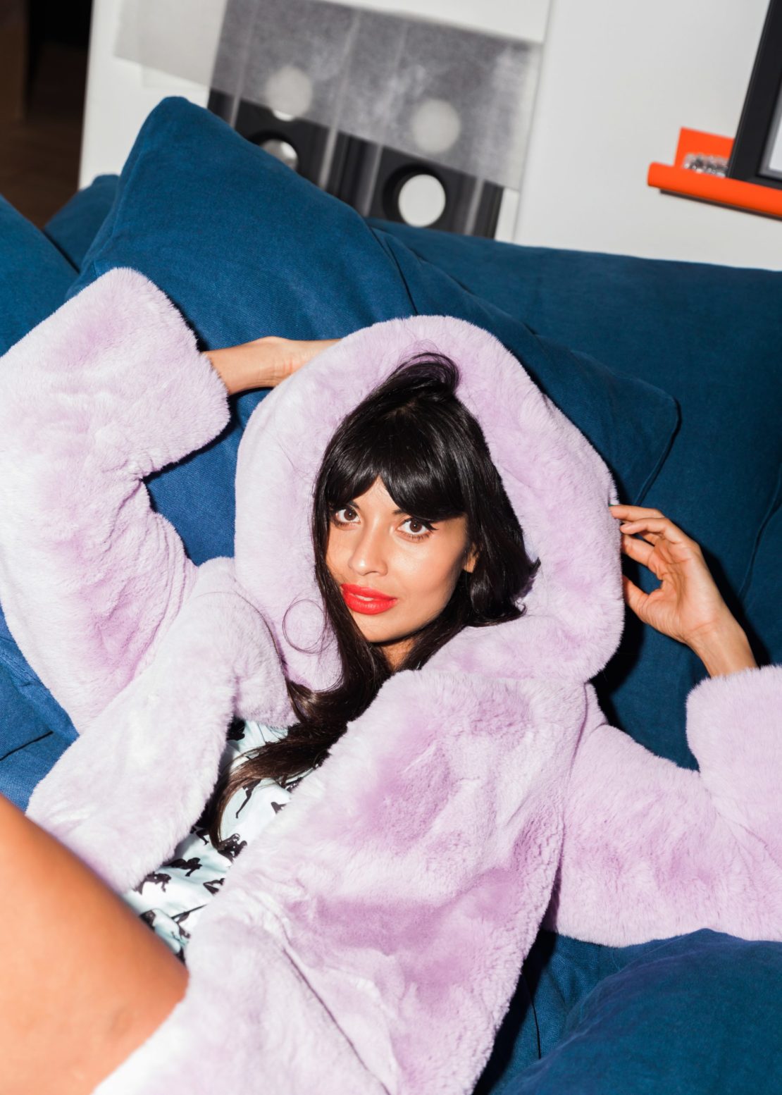 Adult woman in purple faux fur hooded jacket sits on a blue sofa with red lipstick and dark hair posing inside a home.