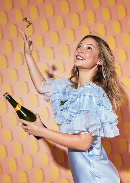 Happy adult female in ruffled dress holds a champagne bottle and throws a golden object against a peach-and-yellow geometric background.