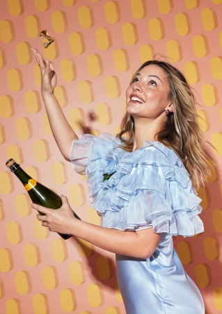 Happy adult female in ruffled dress holds a champagne bottle and throws a golden object against a peach-and-yellow geometric background.