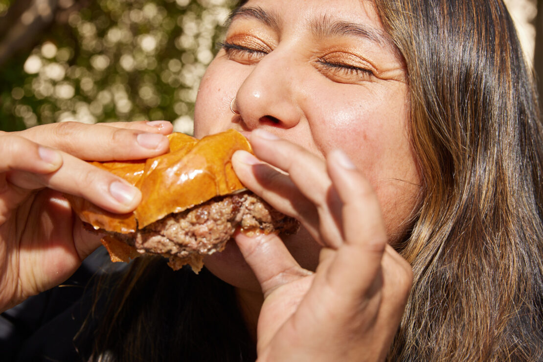 Woman with closed eyes biting into a burger with visible meat and glazed bun, outdoors with blurred tree background, close-up.