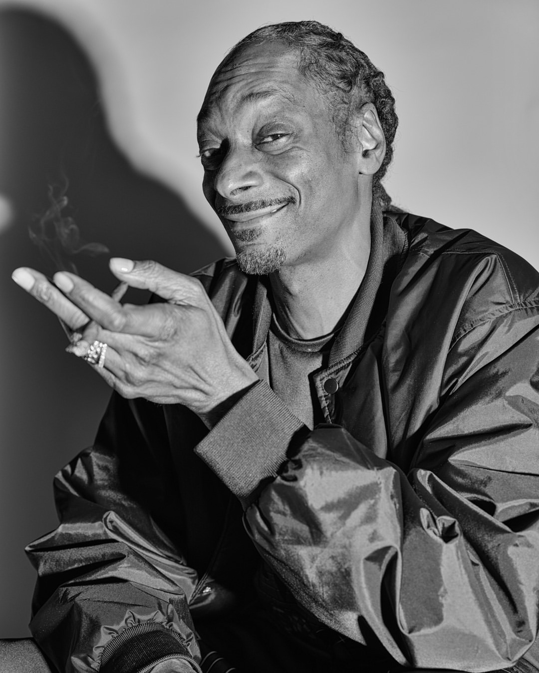 Black and white studio portrait of Snoop Dogg, smoking, wearing a jacket and rings, smiling slightly, with a shadow background.