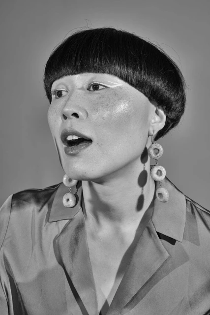 Monochromatic portrait of a woman with a blunt bob haircut and white eyeliner, wearing donut earrings and a collared shirt.
