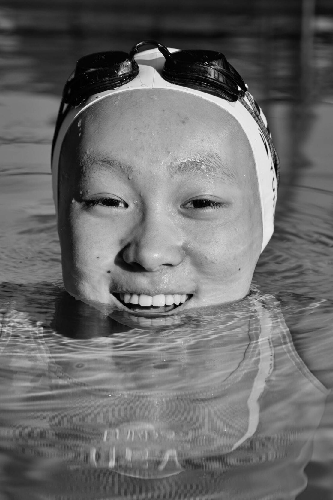 Close-up black and white portrait of an Asian female swimmer in a pool, wearing a swim cap and goggles on her head, smiling at the camera.
