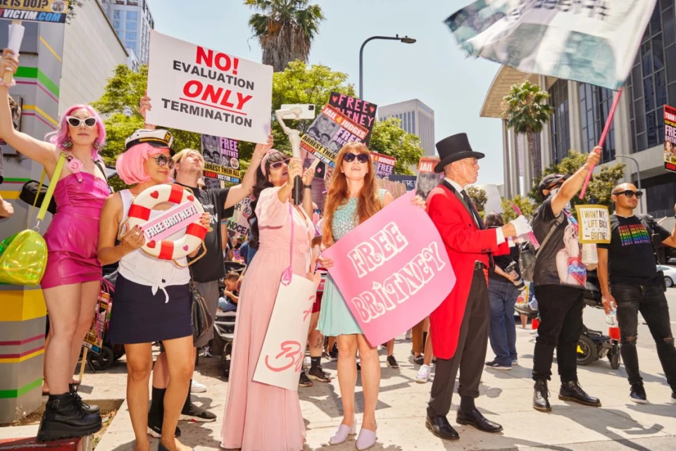 Outdoor daytime shot of a "Free Britney" protest; a crowd of people is holding signs on a city street with buildings and a sunny sky.