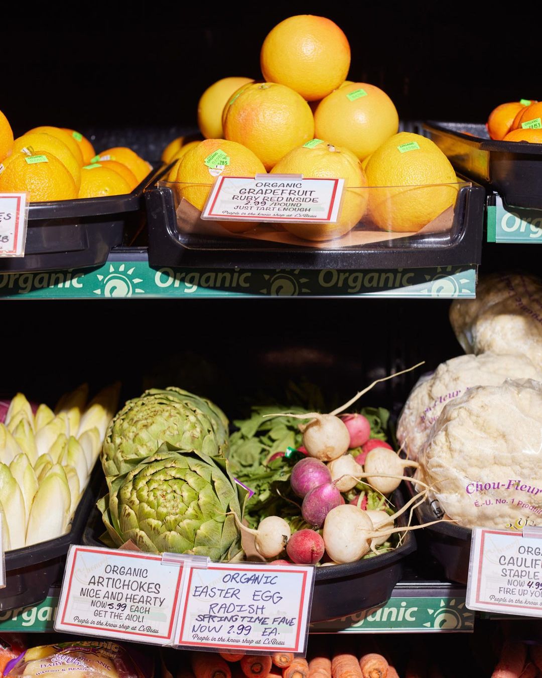 Assortment of fresh produce including grapefruits, artichokes, radishes, and cauliflower on display at a grocery store, organic labels visible.