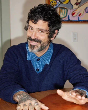 Portrait of a man with dark curly hair and beard wearing a navy blue v-neck sweater over a blue patterned collared shirt.