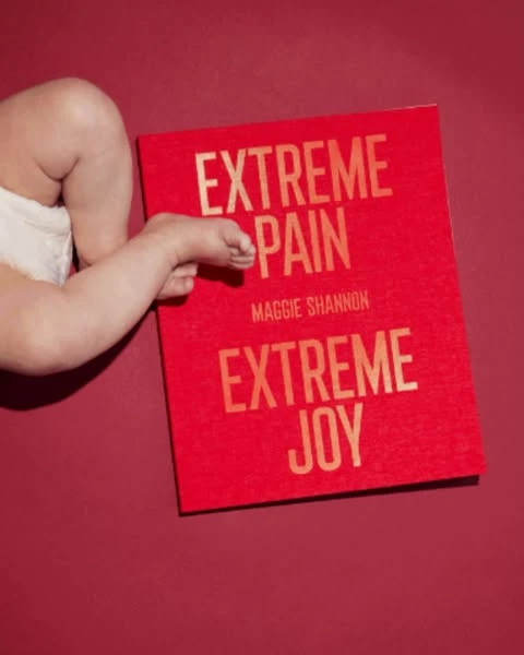 Baby's leg and foot rest on a bright red book titled 'Extreme Pain Extreme Joy' by Maggie Shannon, creating a compelling visual.