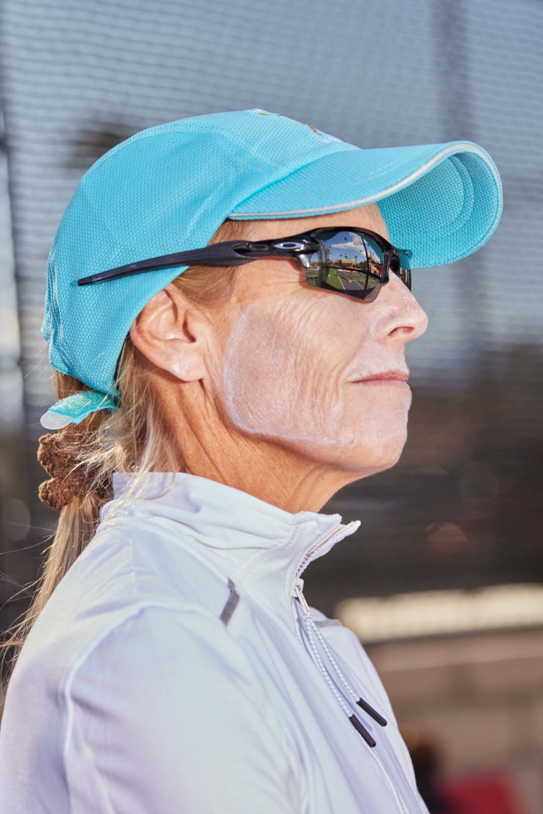 Profile view of a mature woman wearing a turquoise baseball cap and black sports sunglasses with white sunscreen on her face.
