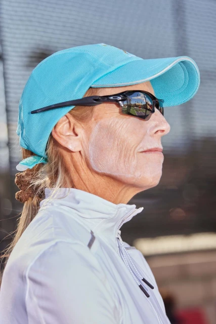 Profile view of a mature woman wearing a turquoise baseball cap and black sports sunglasses with white sunscreen on her face.